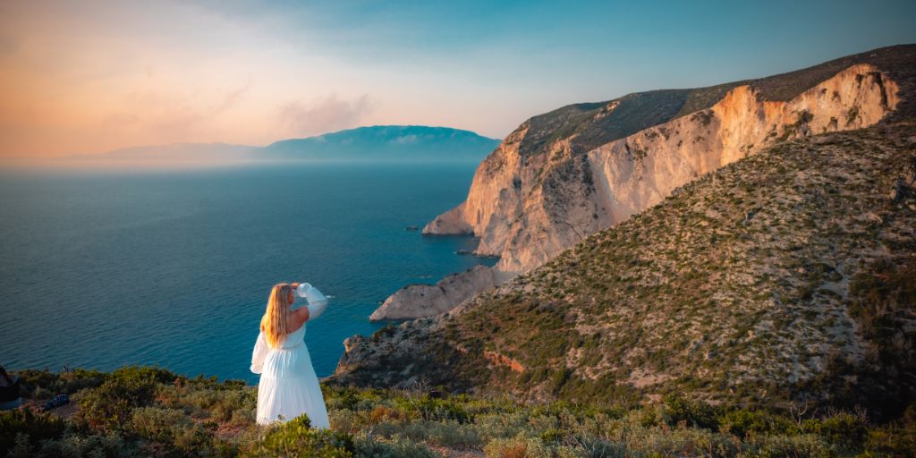 Woman in a flowing white dress stands on the edge of a cliff overlooking beautiful water in Zakynthos Greece