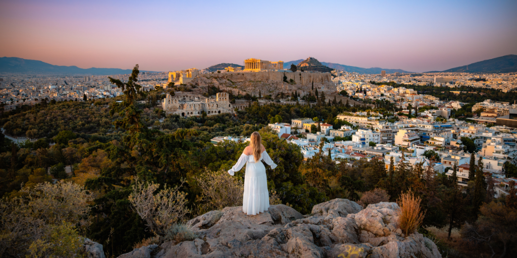 Woman in a flowing white dress stands overlooking Athens greece, with historic landmarks in the background