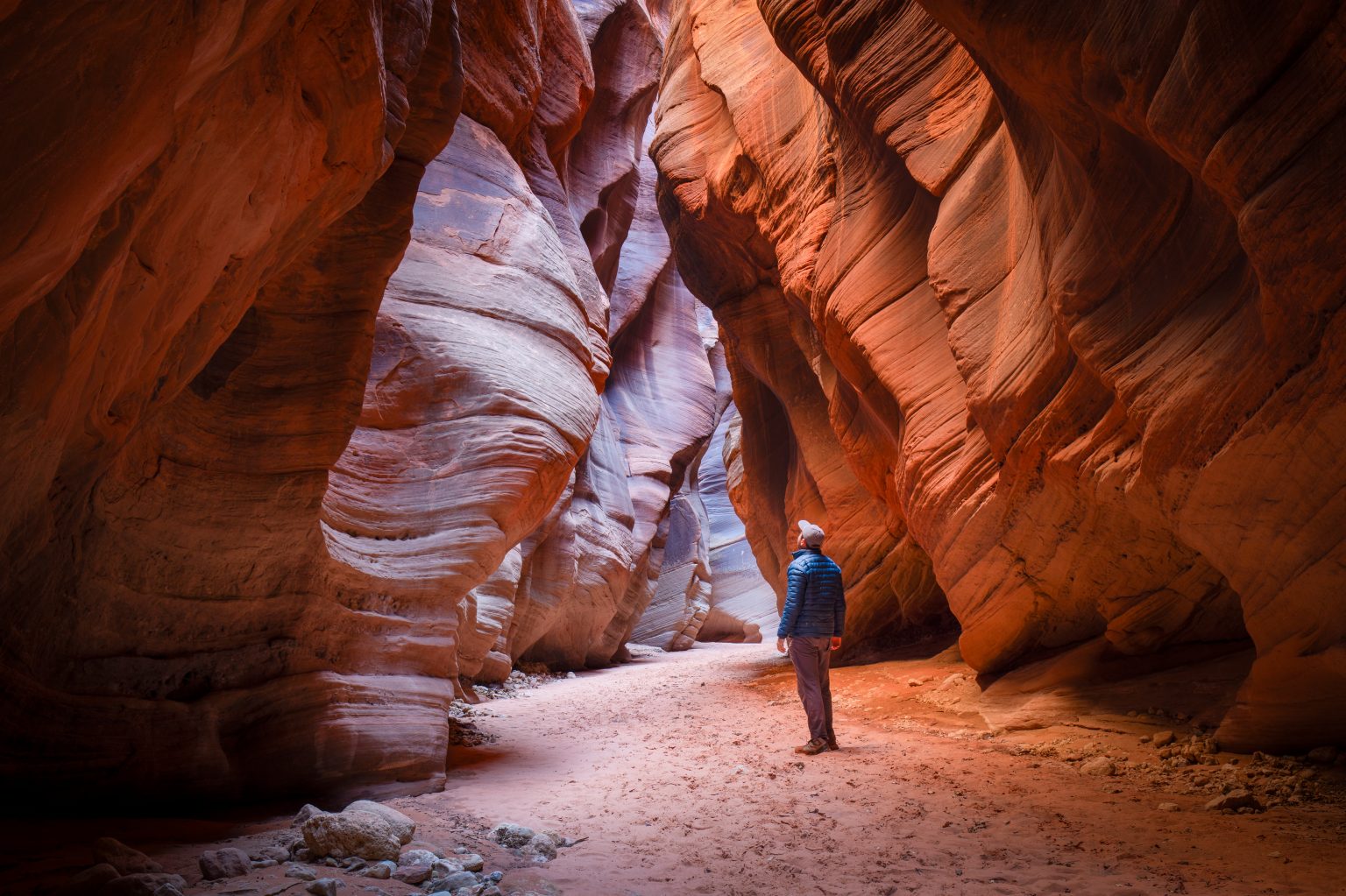 A person in a blue jacket stands inside a narrow red sandstone slot canyon, looking up at the towering, curved rock walls illuminated by soft natural light.