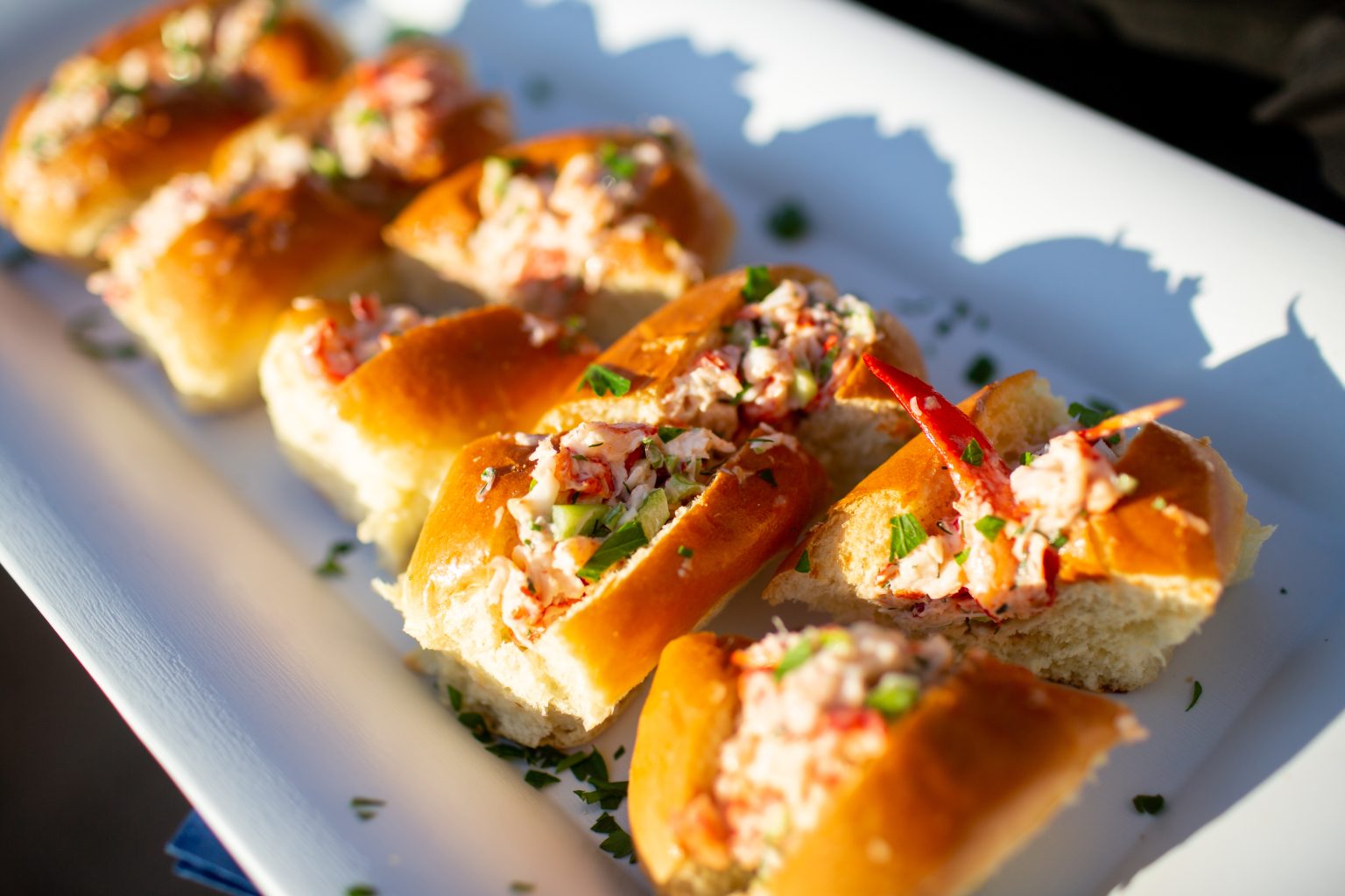 Close-up of mini lobster rolls served on a white rectangular plate, featuring soft golden buns filled with lobster salad, garnished with chopped herbs, with one piece topped by a small red lobster claw.