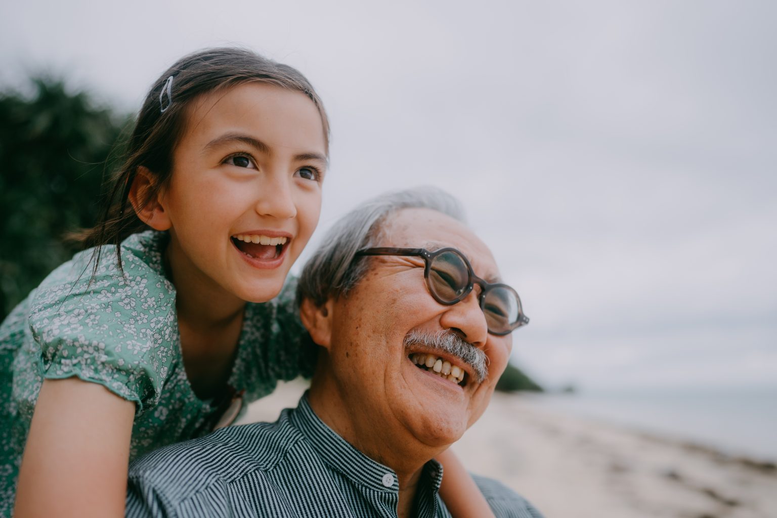 A smiling young girl leans over an older man’s shoulders at the beach, both looking ahead with joyful expressions as they enjoy time together by the water.