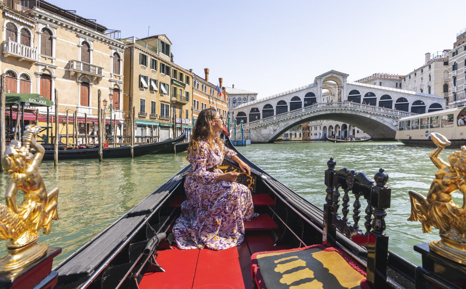 A woman sits on a gondola riding through a canal in Venice, looking toward the Rialto Bridge, with historic buildings lining the water and boats passing nearby.