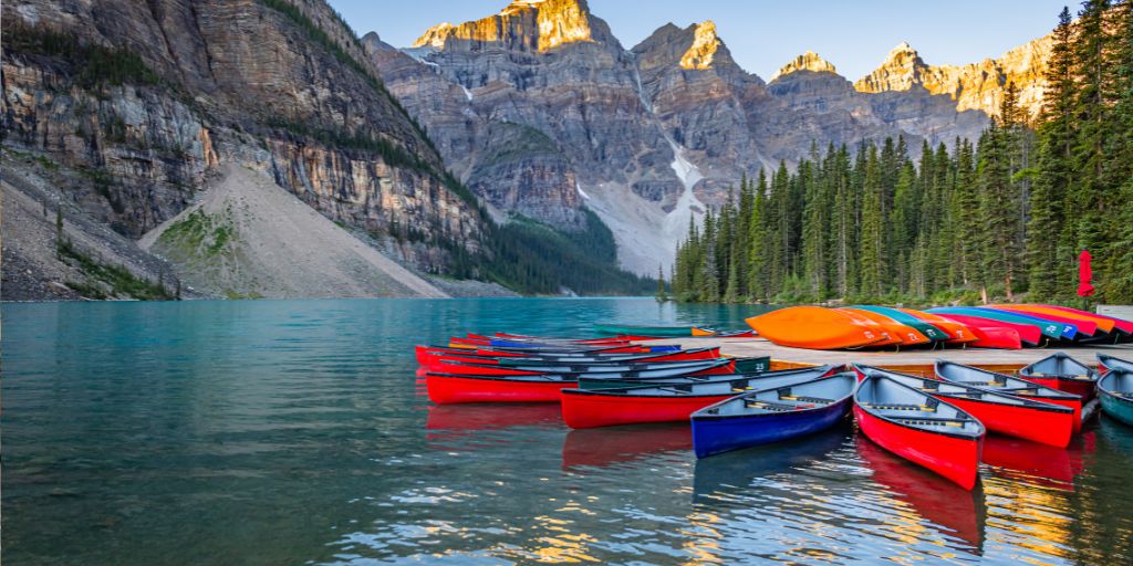 Colorful canoes lined up on a mountain lake with turquoise water, surrounded by pine trees and dramatic rocky peaks glowing in the sunlight.