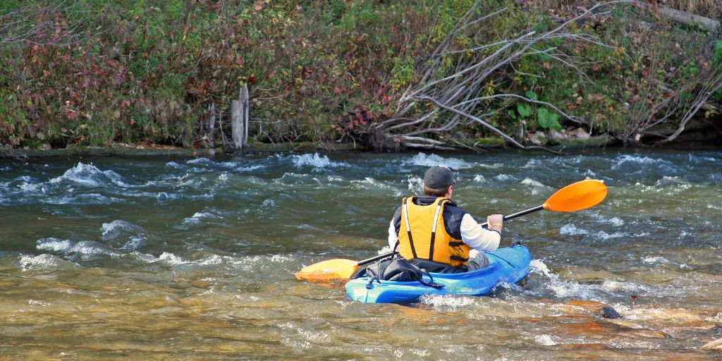 Person kayaking through a shallow, fast-moving river in a blue kayak, wearing a yellow life vest and using an orange paddle, with a wooded riverbank in the background.