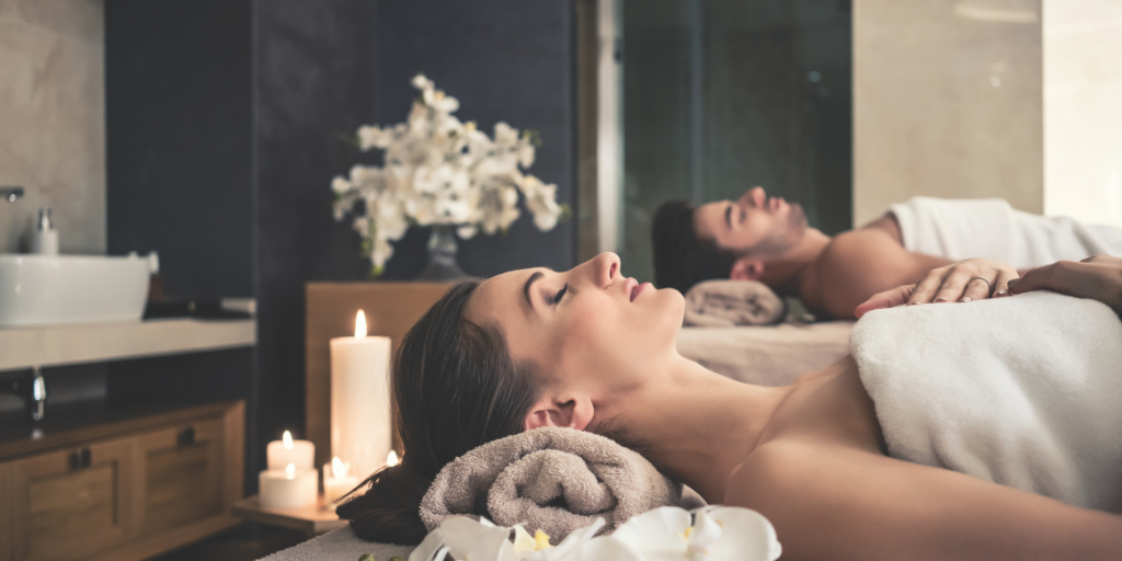 A couple relaxes side by side on massage tables during a spa treatment, surrounded by soft candlelight and rolled towels. The tranquil setting, accented by white flowers and warm lighting, creates a soothing atmosphere for rest and rejuvenation.