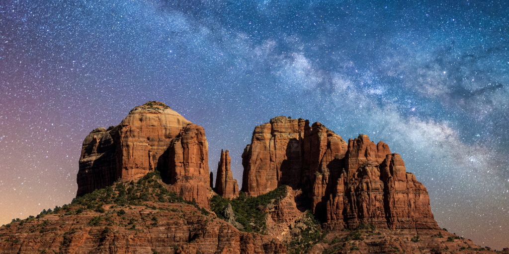 Red rock formations rise beneath a star-filled night sky, with the Milky Way stretching above the desert landscape.