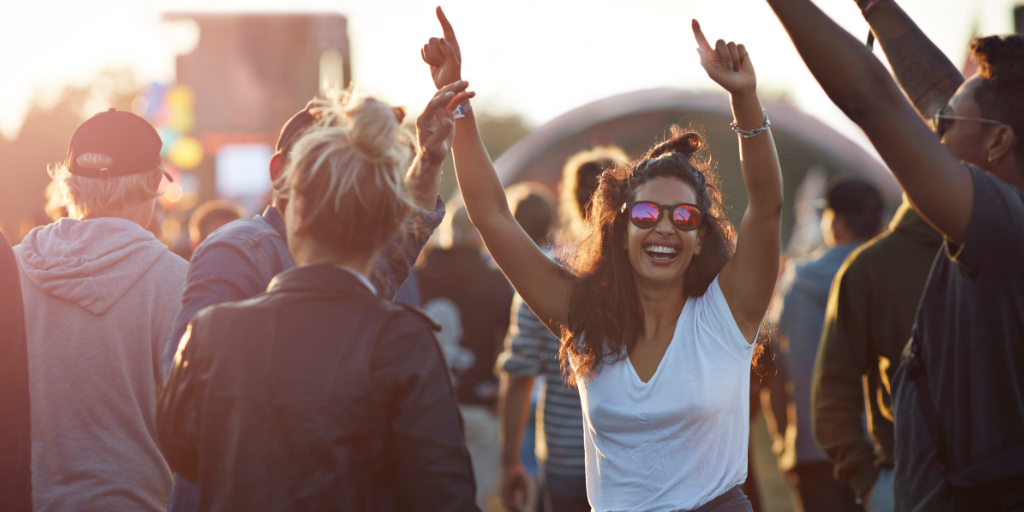 Smiling woman in sunglasses raises her arms while dancing in a crowd at an outdoor event during sunset.
