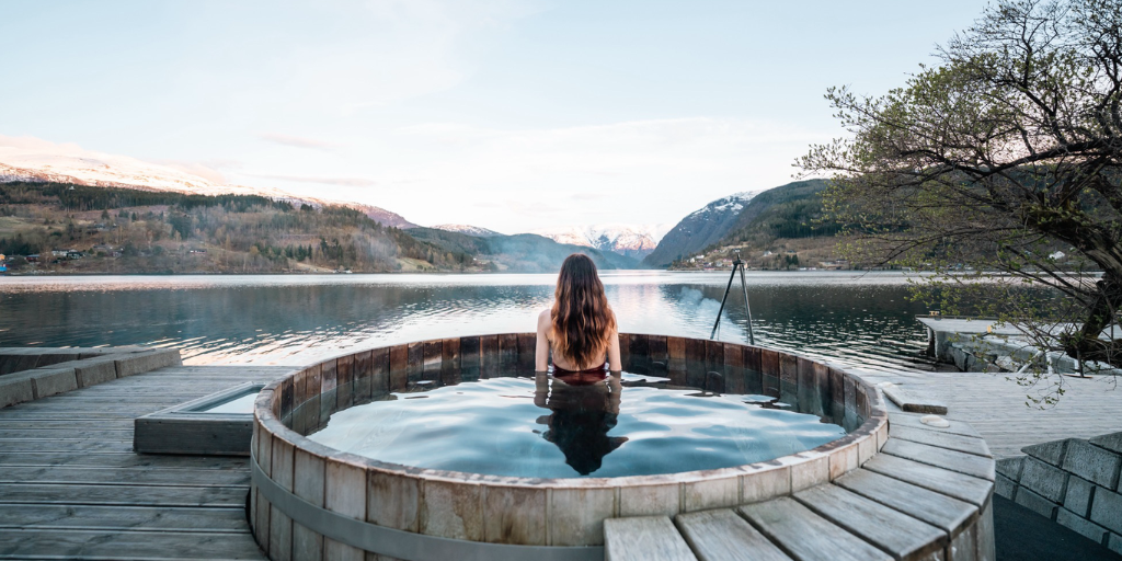 Woman relaxes in a round wooden hot tub overlooking a calm lake surrounded by snowcapped mountains.